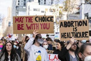 A group of protestors holding signs that say, “May the choice be with you” and “abortion is healthcare.”